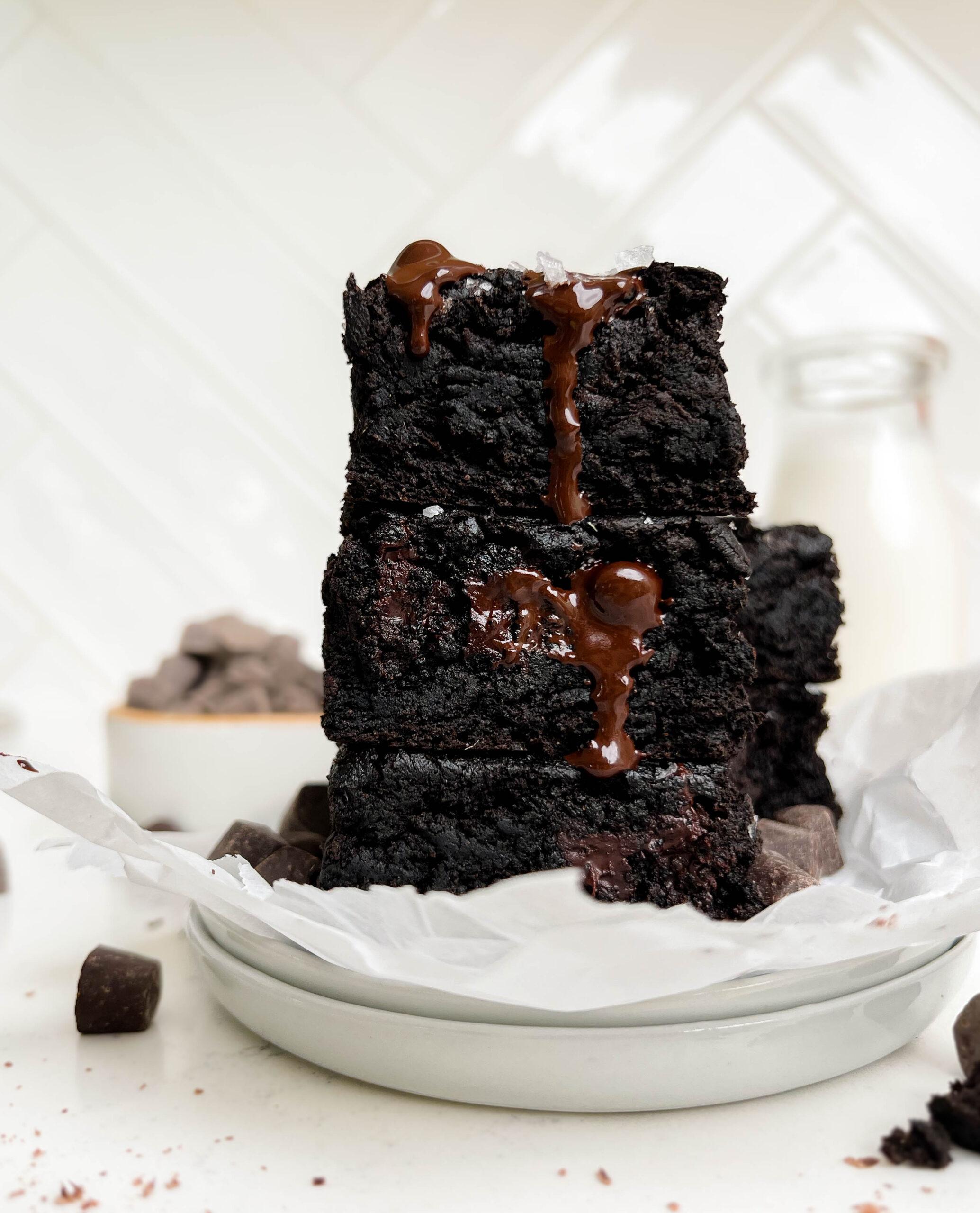 vegan brownies stacked on a white plate next to a bottle of milk and a bowl of chocolate chips
