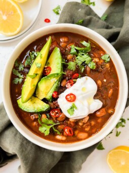 chili in a bowl with avocado, yogurt and spices on top next to a green cloth and lemons