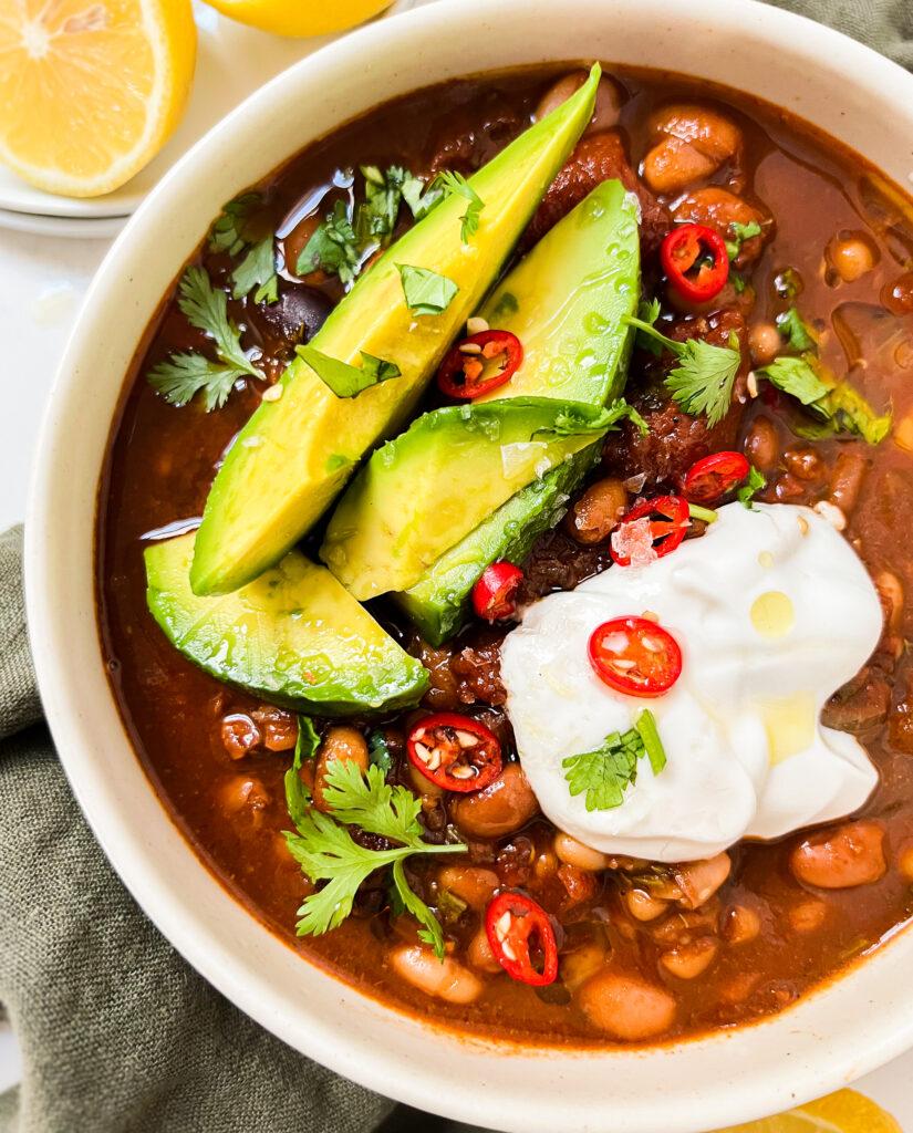 chili in a bowl with avocado, yogurt and spices on top next to a green cloth and lemons