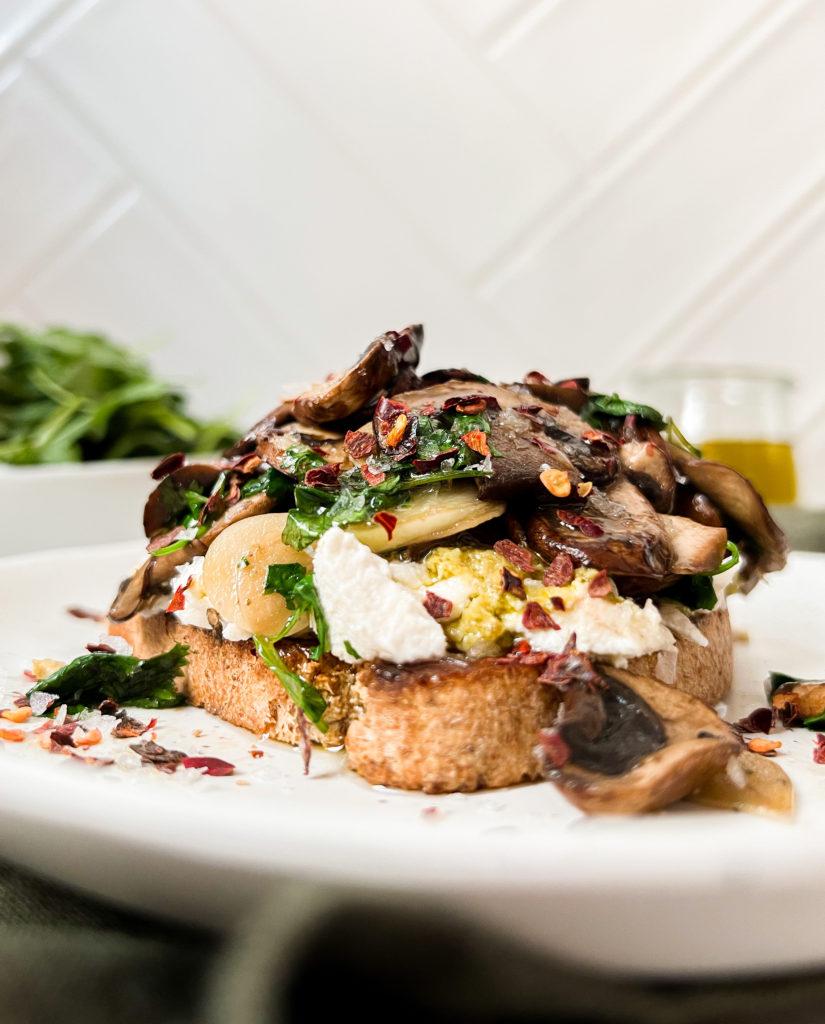 white plate with mushroom toast on top next to a bowl of arugula