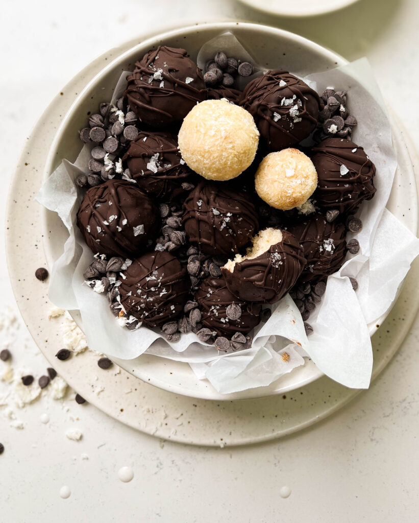 bounty balls in a white bowl next to a glass of milk and a bowl of shredded coconut