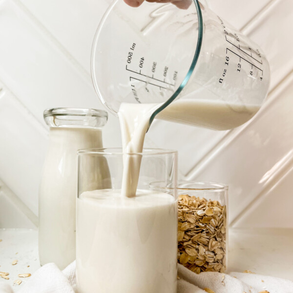 oat milk pouring into a cup next to a jar of milk and a cup of rolled oats