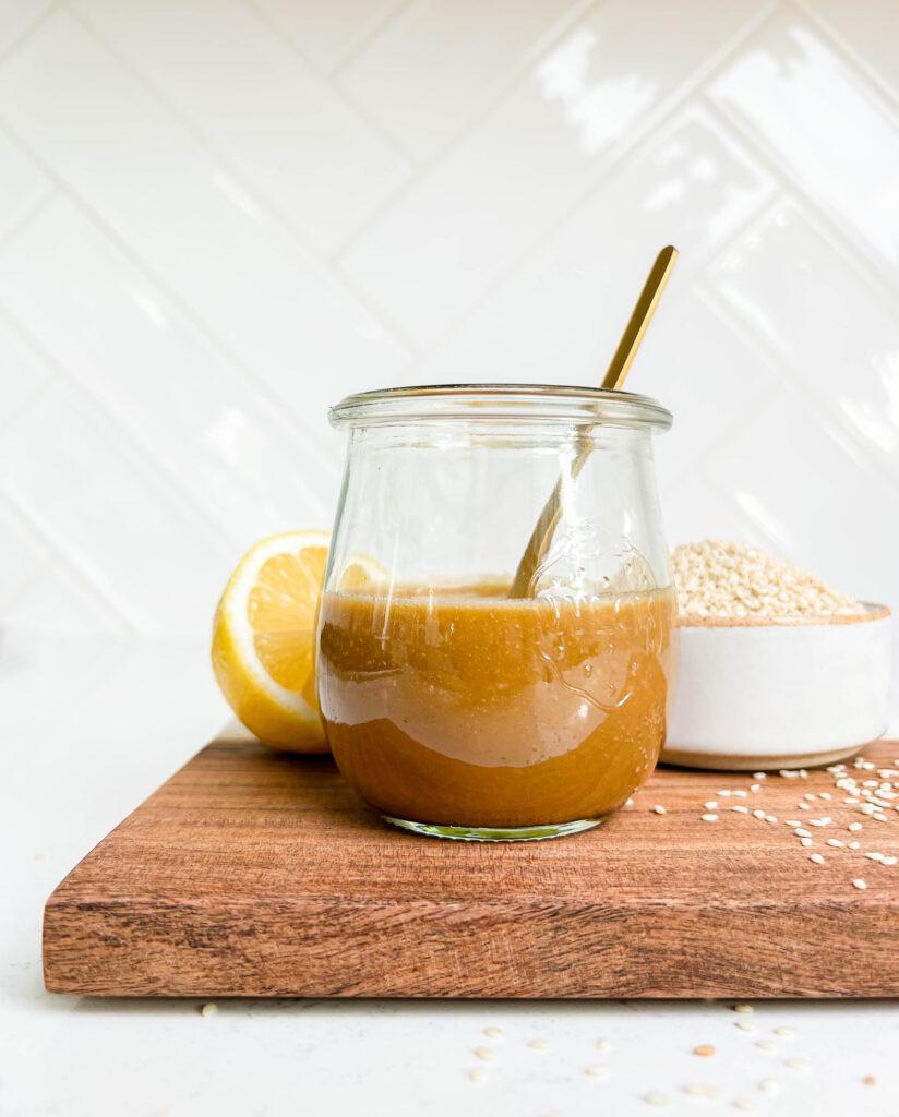 peanut dressing in a glass jar on a cutting board next to lemons and a bowl of sesame seeds