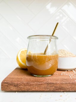 peanut dressing in a glass jar on a cutting board next to lemons and a bowl of sesame seeds