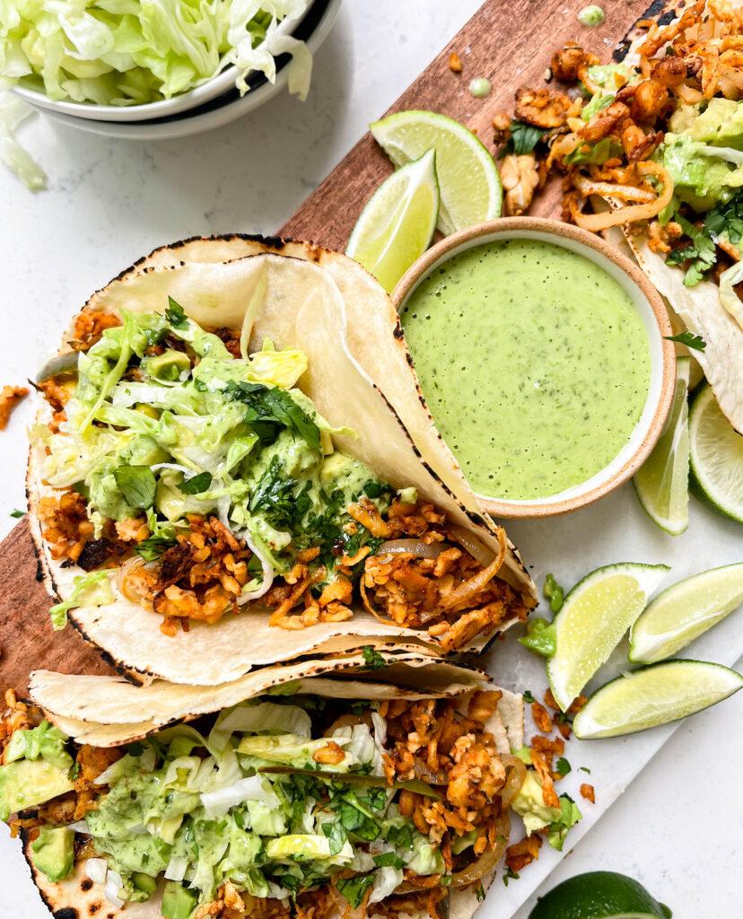 spicy tempeh tacos with a creamy jalapeno verde sauce on a cutting board next to a bowl of lettuce and limes