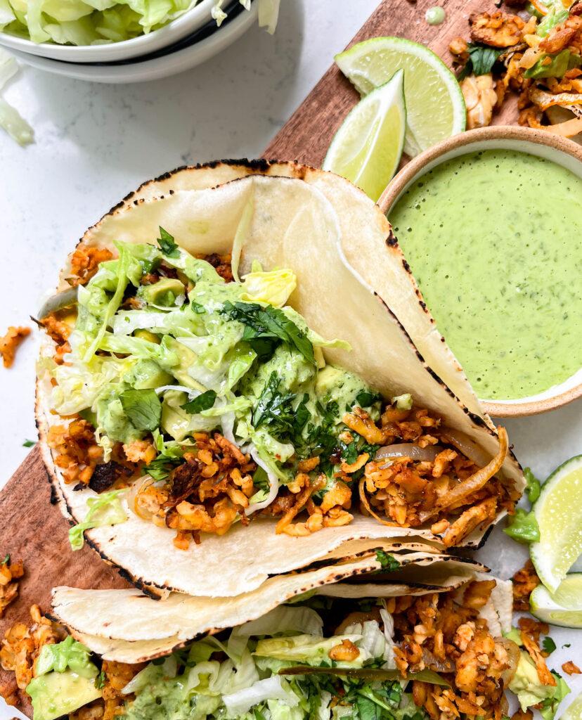 spicy tempeh tacos with a creamy jalapeno verde sauce on a cutting board next to a bowl of lettuce and limes