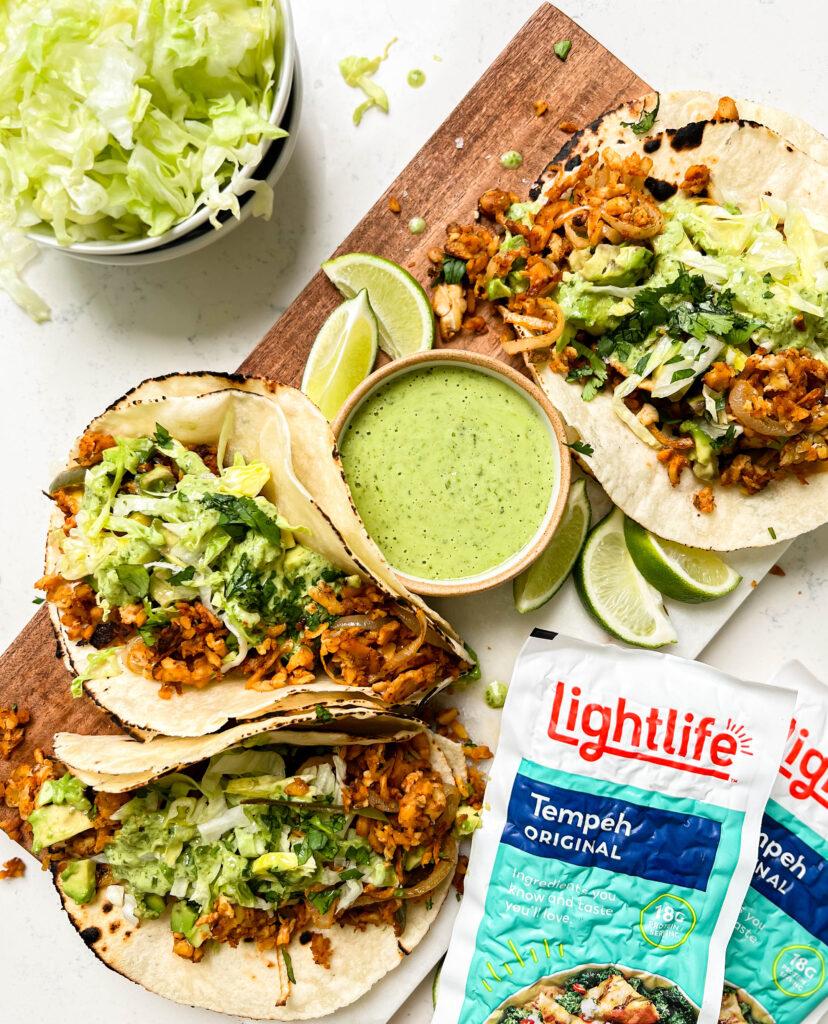 spicy tempeh tacos with a creamy jalapeno verde sauce on a cutting board next to a bowl of lettuce and limes