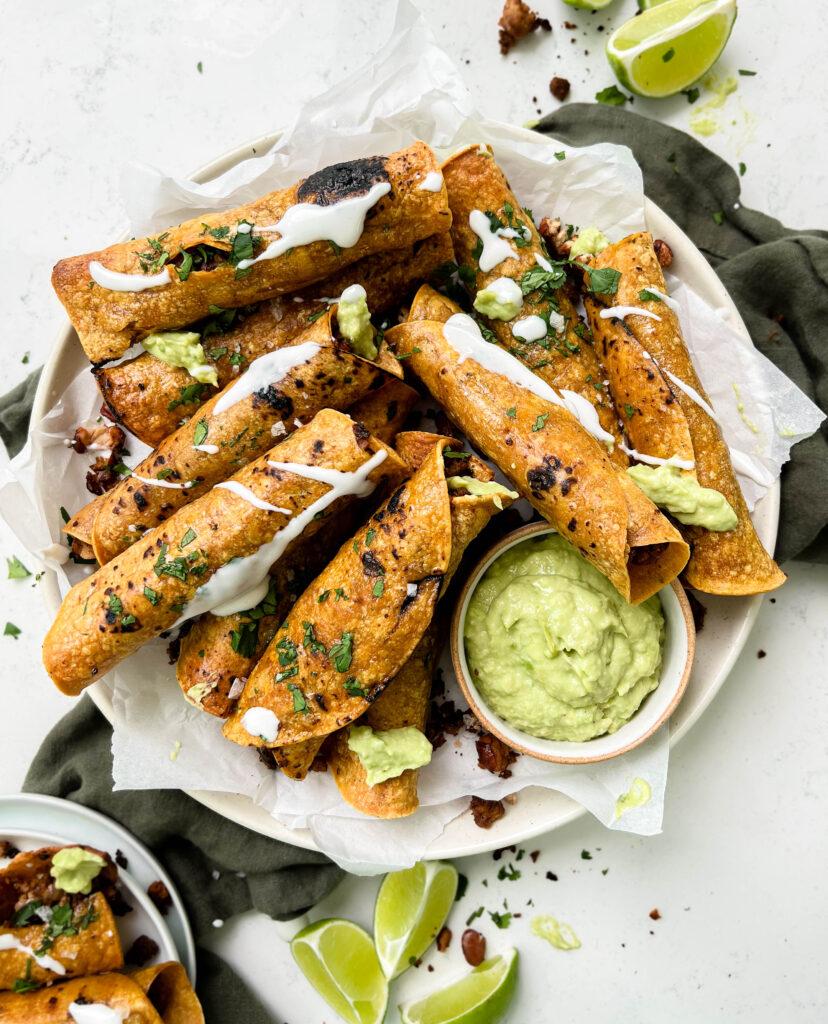 crispy baked vegan taquitos next to avocado crema sauce