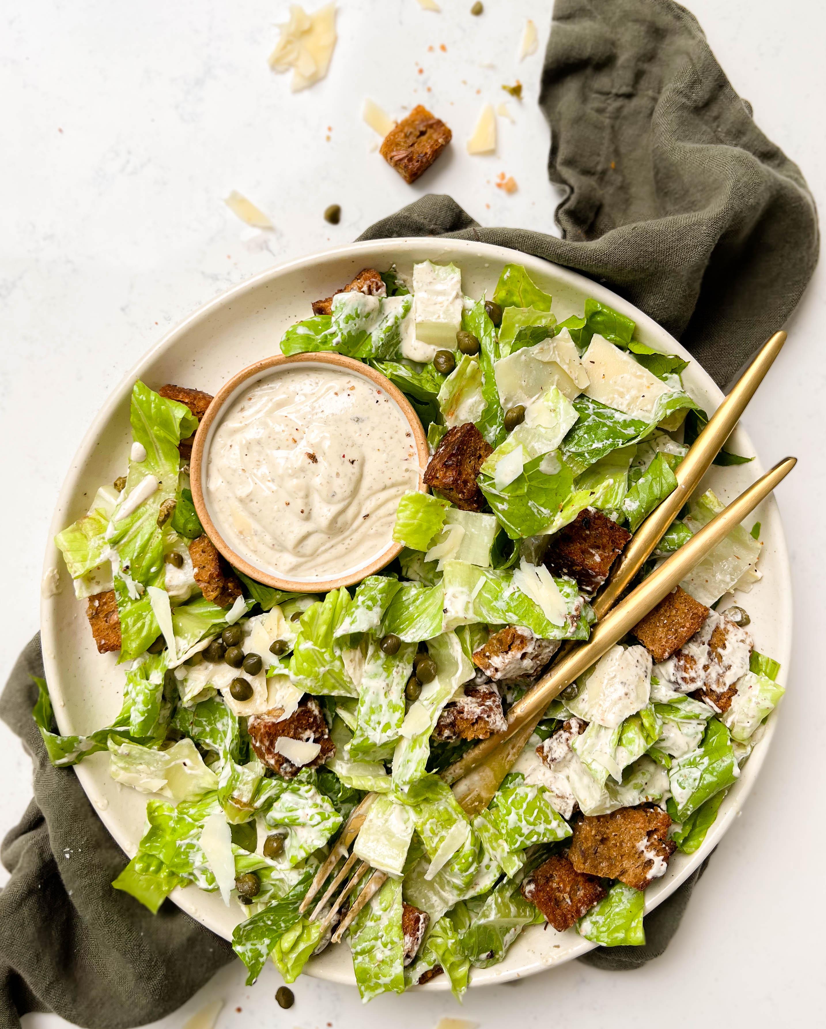 a beige plate with vegan caesar salad, two gold forks and a bowl of dressing on top