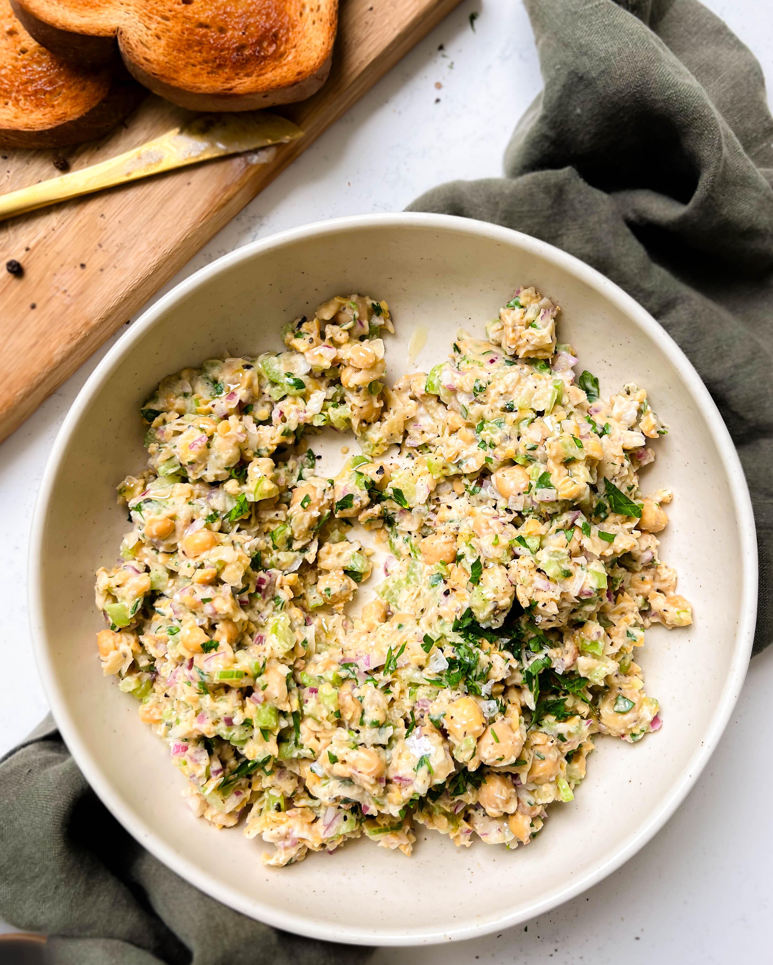 bowl of chickpea tuna salad next to a cutting board with bread and a gold knife