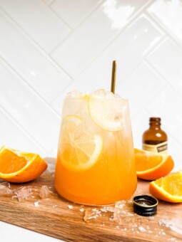a glass of orange spritz on a wooden cutting board next to sliced oranges and vanilla extract