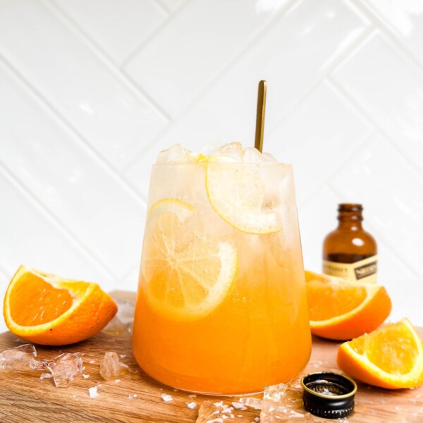 a glass of orange spritz on a wooden cutting board next to sliced oranges and vanilla extract